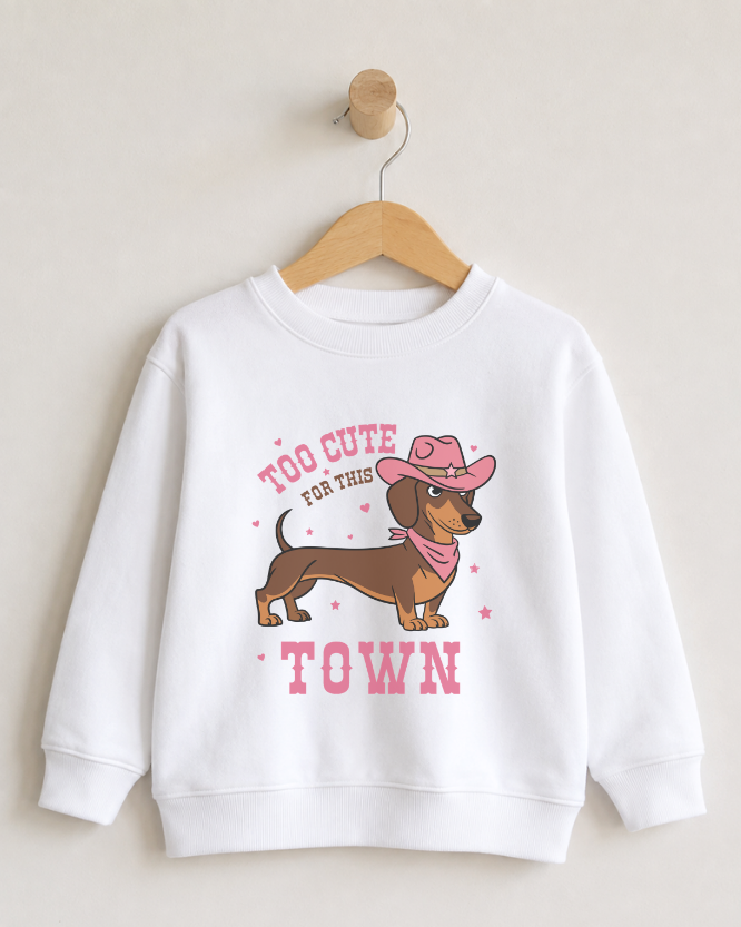 A girls white crewneck sweatshirt hanging on a wooden hanger against a light beige wall. The sweatshirt features a cartoon graphic of a brown dachshund wearing a pink cowboy hat and bandana, with the text 'TOO CUTE FOR THIS TOWN' in pink.