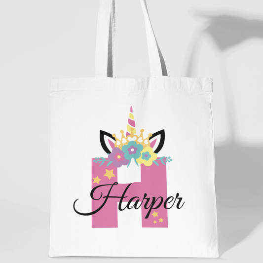 White cotton tote bag featuring a pink letter “H” with a unicorn horn, pastel flowers, and black-tipped ears, personalised with the name “Harper” in elegant black script.