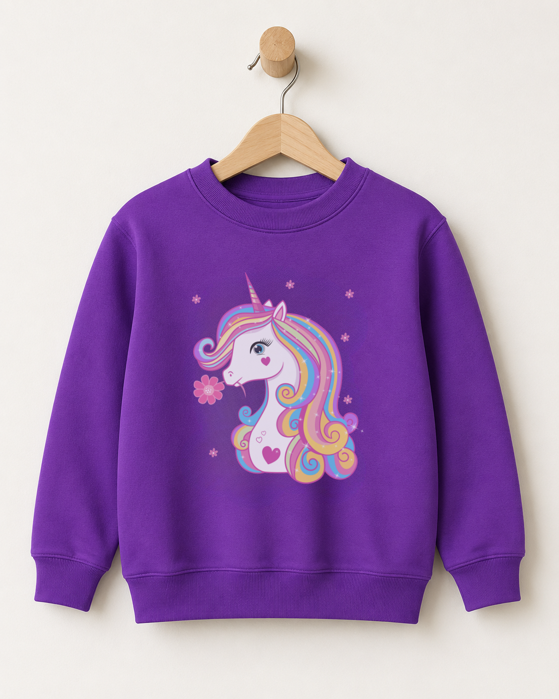 A girls purple crewneck sweatshirt featuring a colourful cartoon unicorn graphic on the front, hanging on a wooden hanger against a plain off-white wall.
