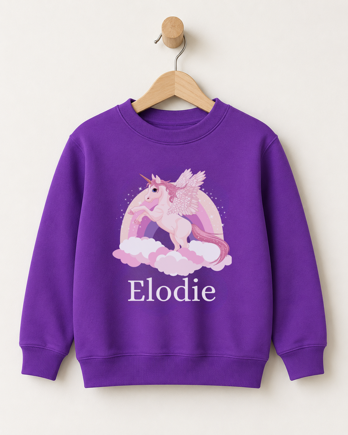 Purple personalised girls’ sweatshirt featuring a white winged unicorn, pastel rainbow, clouds and the name Elodie, displayed on a wooden hanger.