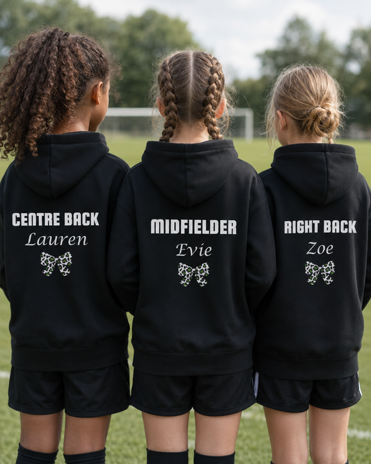 Three girls seen from behind wearing matching black custom-printed athletic hoodies on a grassy sports field. The back of each hoodie features white text naming their position and name ('CENTRE BACK Lauren', 'MIDFIELDER Evie', 'RIGHT BACK Zoe') above a green and white bow graphic.