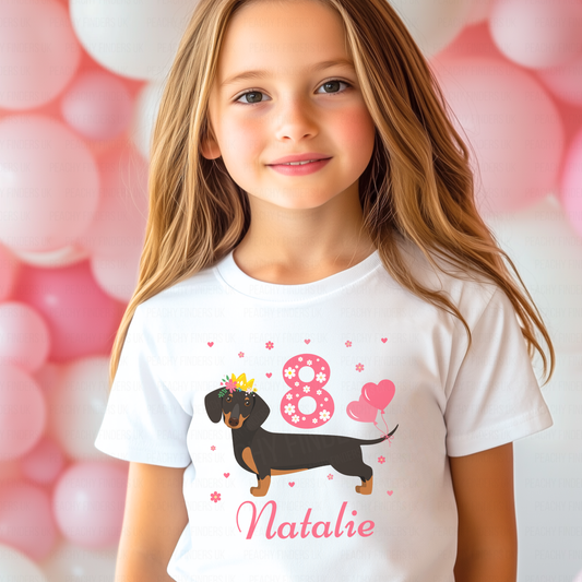 A girl wearing a white t-shirt with a dachshund design and personalised text that reads 'Natalie'. The t-shirt also features numbers '8' with pink hearts and flowers.