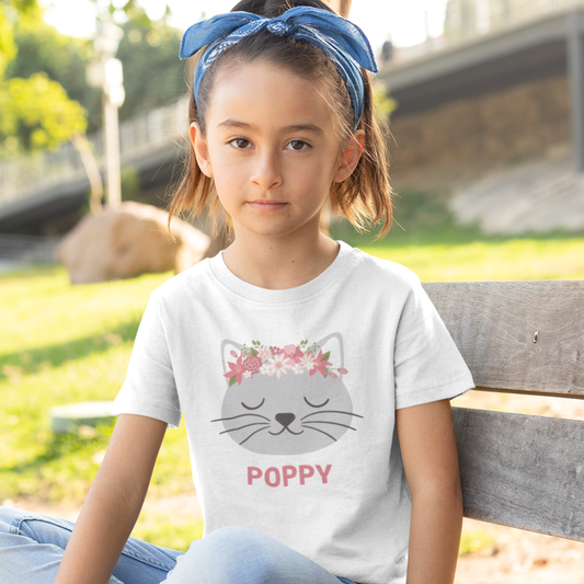 Girl on a park bench wearing white personalised floral cat t-shirt with custom name ‘Poppy’ printed under a grey kitten face with pink flower crown