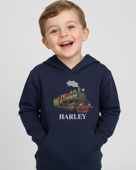 Happy young boy wearing a navy personalised kids hoodie with a vintage steam train graphic and the name Harley, smiling with arms raised against a light studio background.