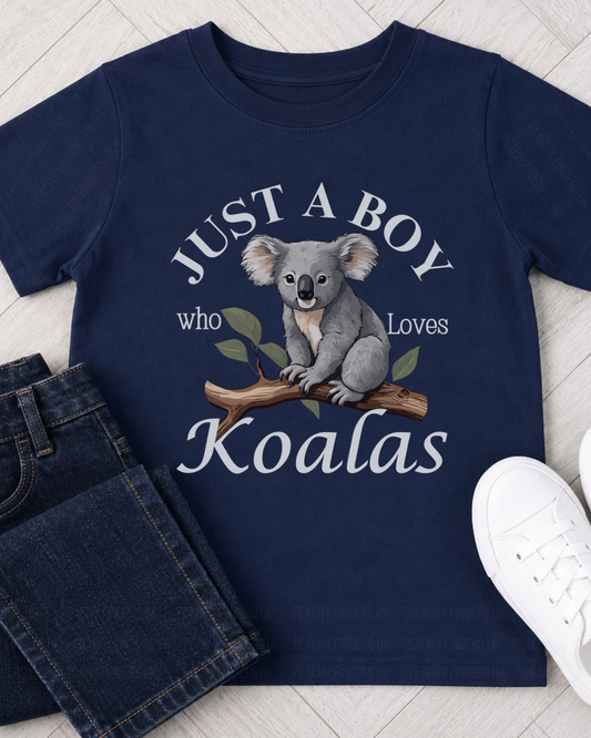 A kids navy blue short-sleeve t-shirt with a graphic of a koala on a branch and the text 'JUST A BOY who Loves Koalas', styled in a flat lay with dark blue jeans and white sneakers on a light wood floor.
