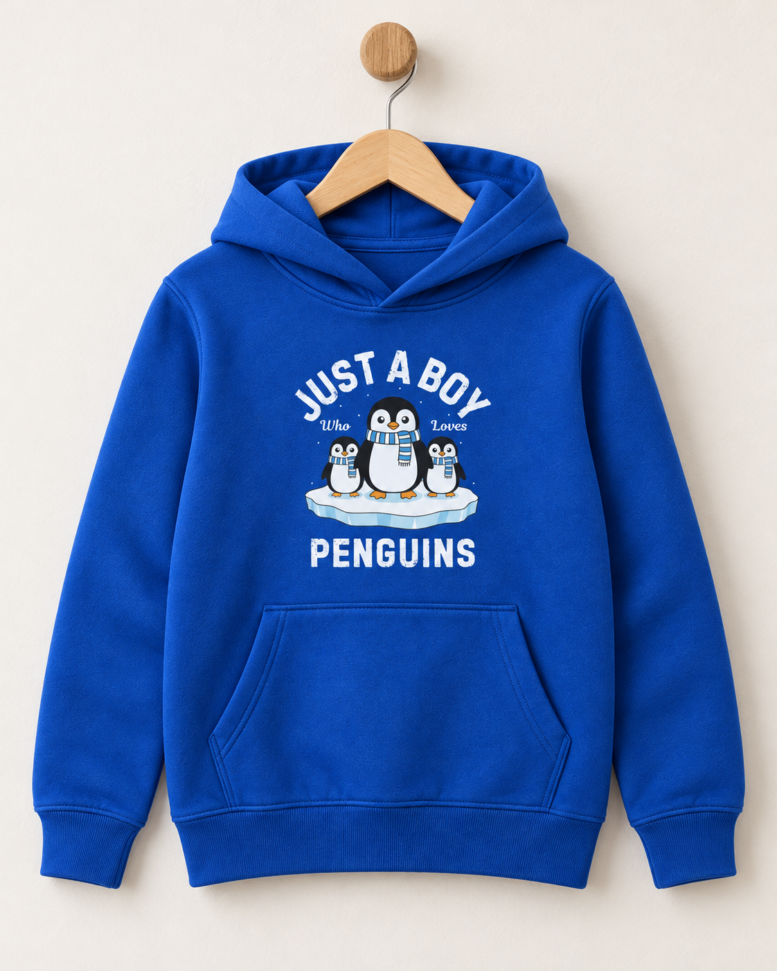 A royal blue kids' hoodie hanging on a wooden hanger against a light beige background. The hoodie features a white graphic print on the front with the text 'JUST A BOY' above a cartoon penguin family and 'PENGUINS' below.