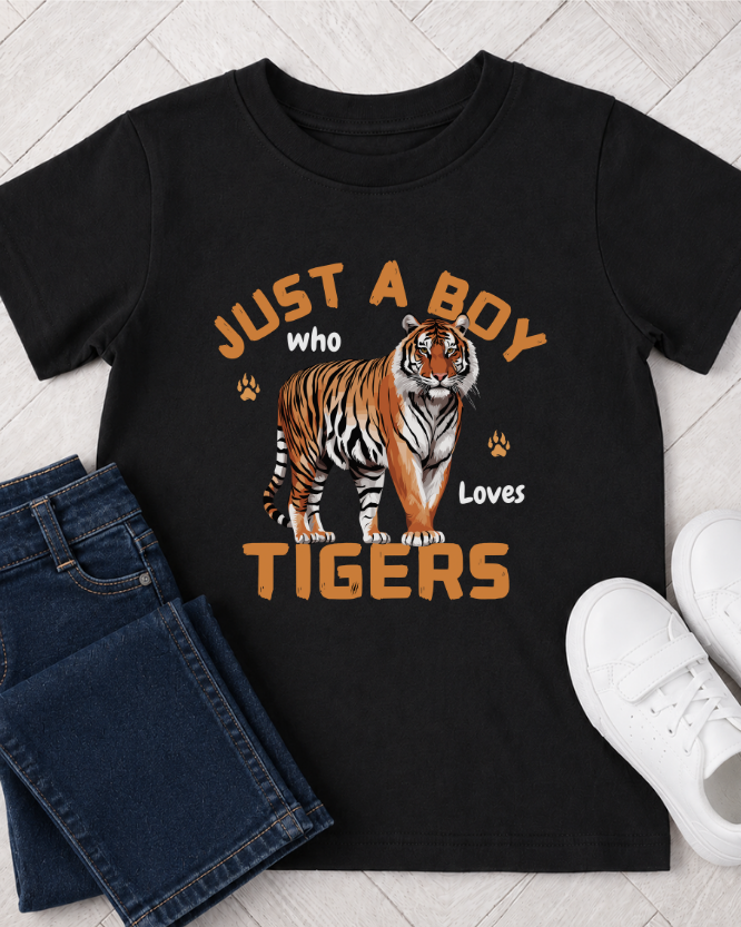 A kids black short-sleeve graphic T-shirt laid flat on a light wood floor, styled with folded blue jeans on the left and white sneakers on the right. The shirt features a large tiger illustration and the text 'JUST A BOY who Loves TIGERS' in orange and white.