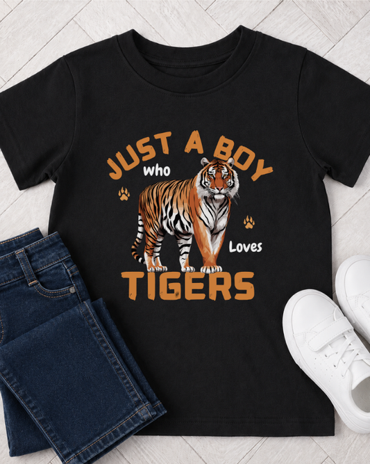 A kids black short-sleeve graphic T-shirt laid flat on a light wood floor, styled with folded blue jeans on the left and white sneakers on the right. The shirt features a large tiger illustration and the text 'JUST A BOY who Loves TIGERS' in orange and white.