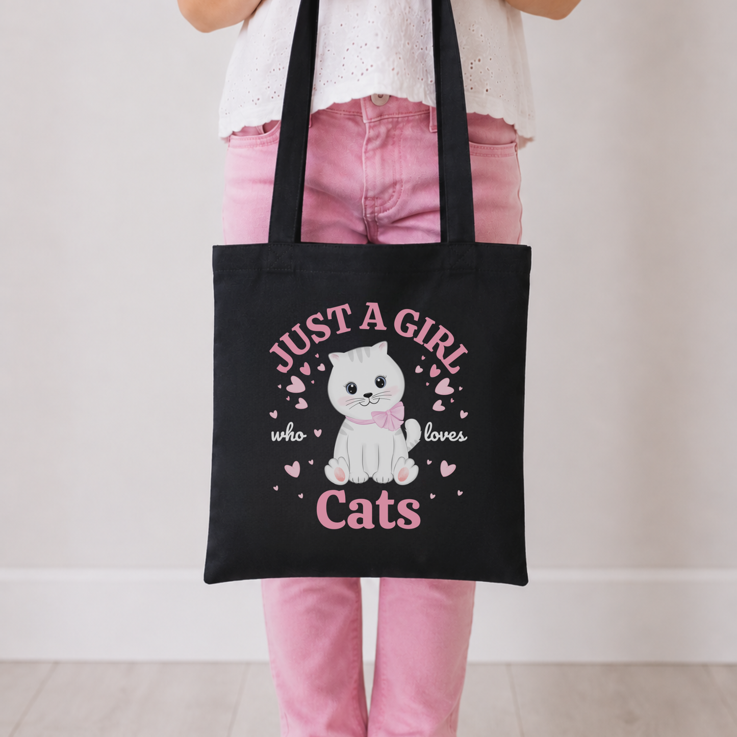 A young girl wearing pink jeans and a white top holds a black cotton tote bag with a white cat illustration and the text 'JUST A GIRL who loves Cats' in pink, against a plain indoor background.