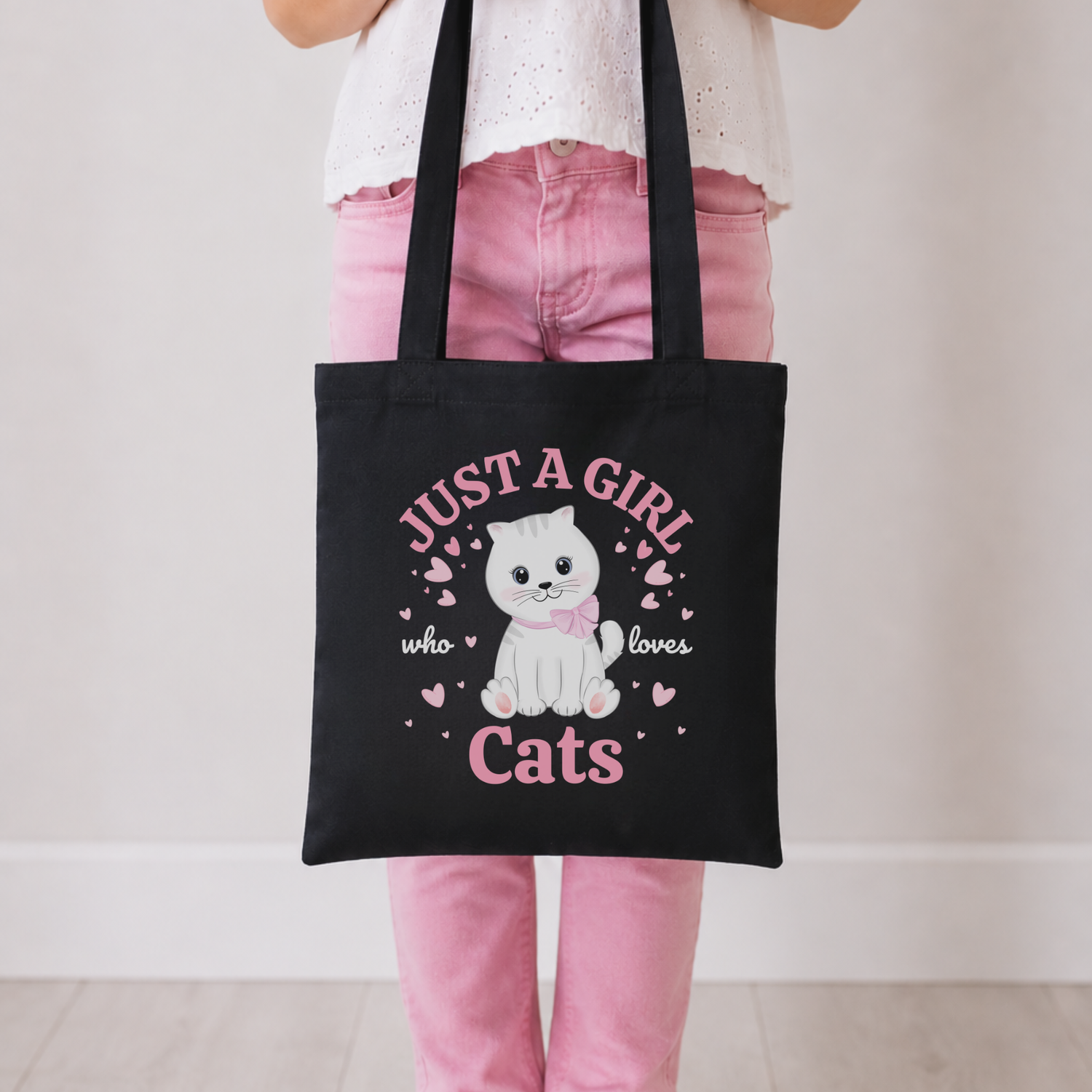 A young girl wearing pink jeans and a white top holds a black cotton tote bag with a white cat illustration and the text 'JUST A GIRL who loves Cats' in pink, against a plain indoor background.