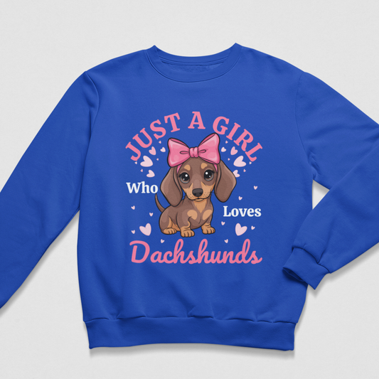 A kids royal blue crewneck sweatshirt featuring a cartoon dachshund wearing a pink bow, surrounded by the text 'JUST A GIRL Who Loves Dachshunds' in pink and white, with small pink hearts.