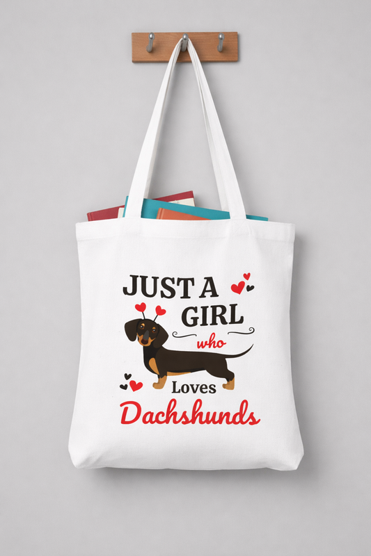 “Just A Girl Who Loves Dachshunds” white tote bag hanging on wooden coat hooks, filled with colourful books, long handles, minimalist grey wall background.