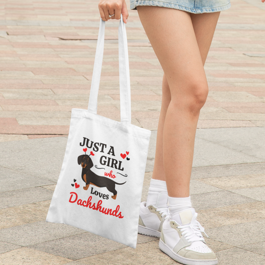 A person holding a white cotton tote bag with the text 'JUST A GIRL WHO LOVES DACHSHunds' and an illustration of a dachshund on it.