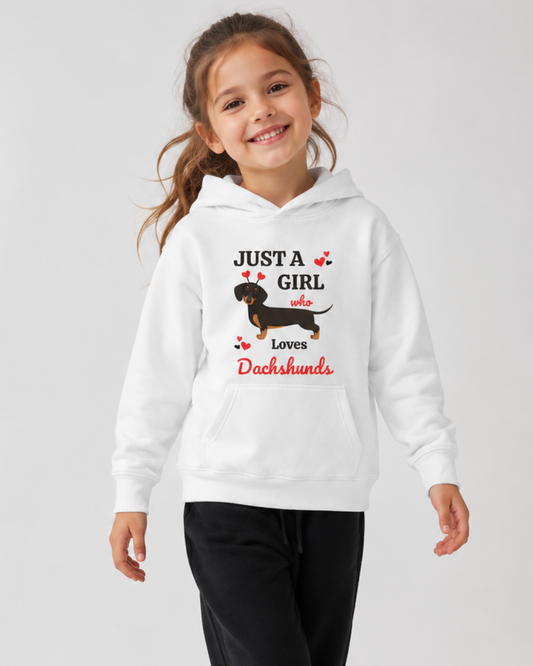 A young girl smiling while wearing a white pullover hoodie with a graphic print that reads 'JUST A GIRL who Loves Dachshunds' with an illustration of a dachshund. She is also wearing black joggers, set against a plain light grey studio background.
