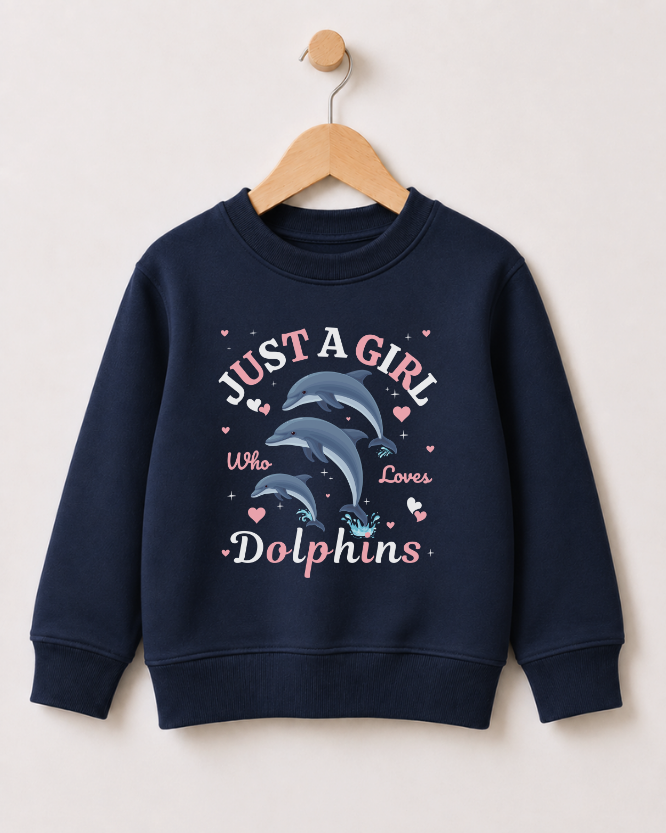 A girls navy blue crewneck sweatshirt hanging on a wooden hanger against a light beige wall. The sweatshirt features a graphic print of two dolphins with the text 'JUST A GIRL Who Loves Dolphins' in pink and white, accented with small pink hearts.