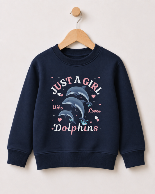 A girls navy blue crewneck sweatshirt hanging on a wooden hanger against a light beige wall. The sweatshirt features a graphic print of two dolphins with the text 'JUST A GIRL Who Loves Dolphins' in pink and white, accented with small pink hearts.