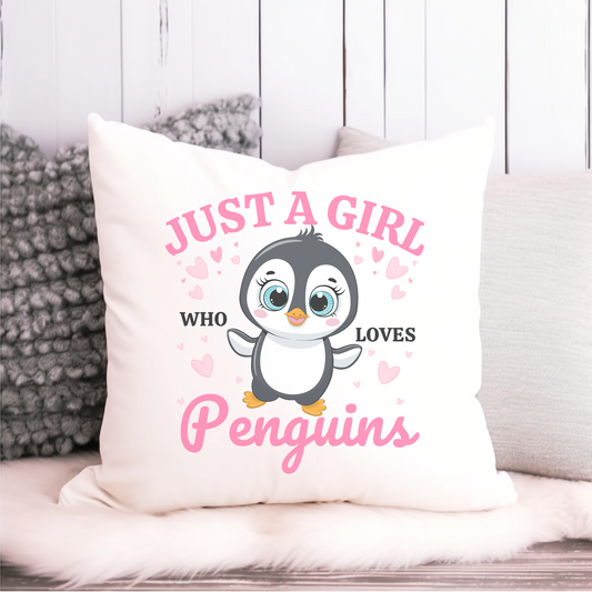A white square throw pillow displays a cute cartoon penguin with big blue eyes and rosy cheeks, surrounded by small pink hearts. Above the penguin, in a slightly curved pink font, are the words "JUST A GIRL". To the left and right of the penguin are the words "WHO" and "LOVES" in a smaller black font. Below the penguin, in a larger, flowing pink script, is the word "Penguins". 