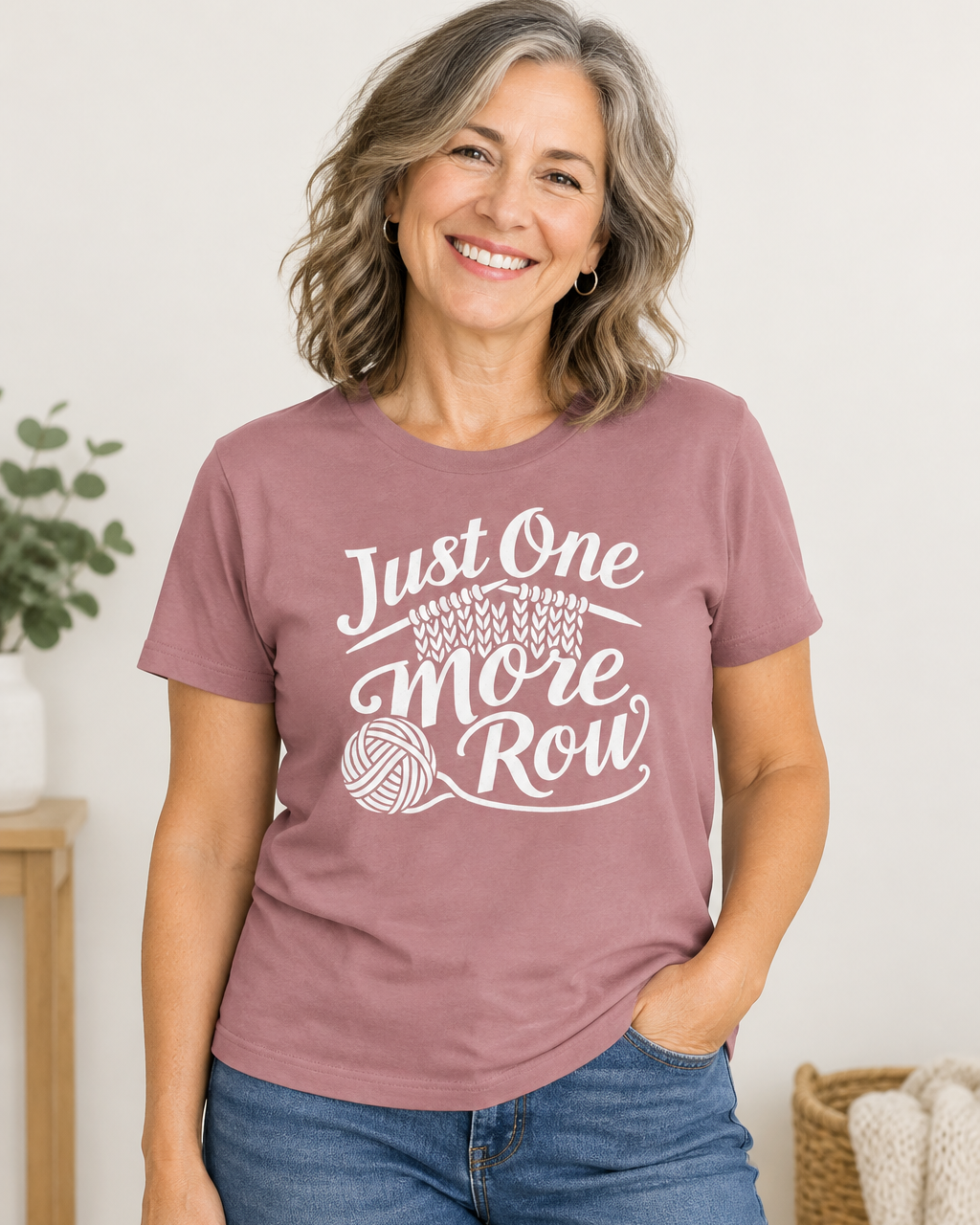 A smiling woman with shoulder-length hair wears a dusty mauve t-shirt featuring a white screen-printed graphic that reads "Just One More Row" with a ball of yarn and knitting needles. She is wearing light blue jeans against a simple off-white background.