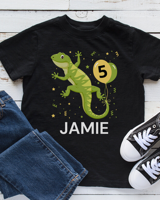 A black children's t-shirt with a colorful illustration of a green gecko and the name 'Jamie' printed on it, accompanied by the number '5'. The t-shirt is displayed flat with a pair of jeans and a pair of sneakers.