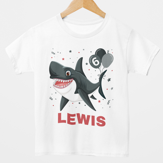 kids white t-shirt on hanger with a birthday themed cartoon shark holding black and grey balloons with the number six, falling confetti surround the shark and name. 