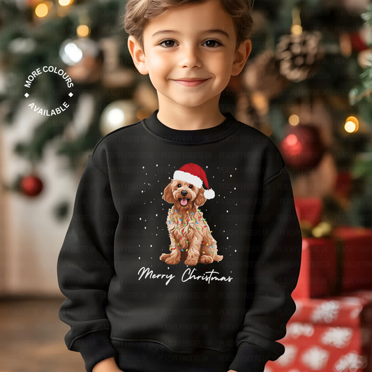 Smiling young boy wearing a black Christmas sweatshirt featuring a cute Cockapoo dog in a Santa hat wrapped in festive lights, standing by a decorated Christmas tree.