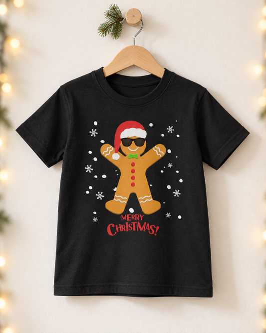 A black short-sleeve kids' t-shirt hanging on a wooden hanger against a warm beige wall with soft holiday string lights. The shirt features a graphic of a gingerbread man wearing a Santa hat and sunglasses, surrounded by white snowflakes, with 'MERRY CHRISTMAS!' printed in red below.