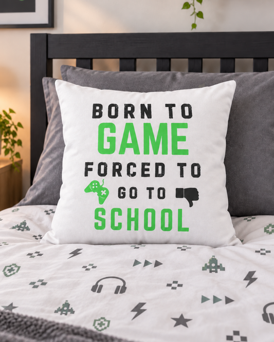 A white square throw pillow with the text 'BORN TO GAME FORCED TO GO TO SCHOOL' in black and green, styled on a bed with a dark wood headboard and matching gaming-themed bedding.