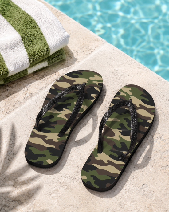 A pair of kids camouflage print flip-flop sandals with black straps, placed on a light stone deck next to a green and white striped towel and a swimming pool.