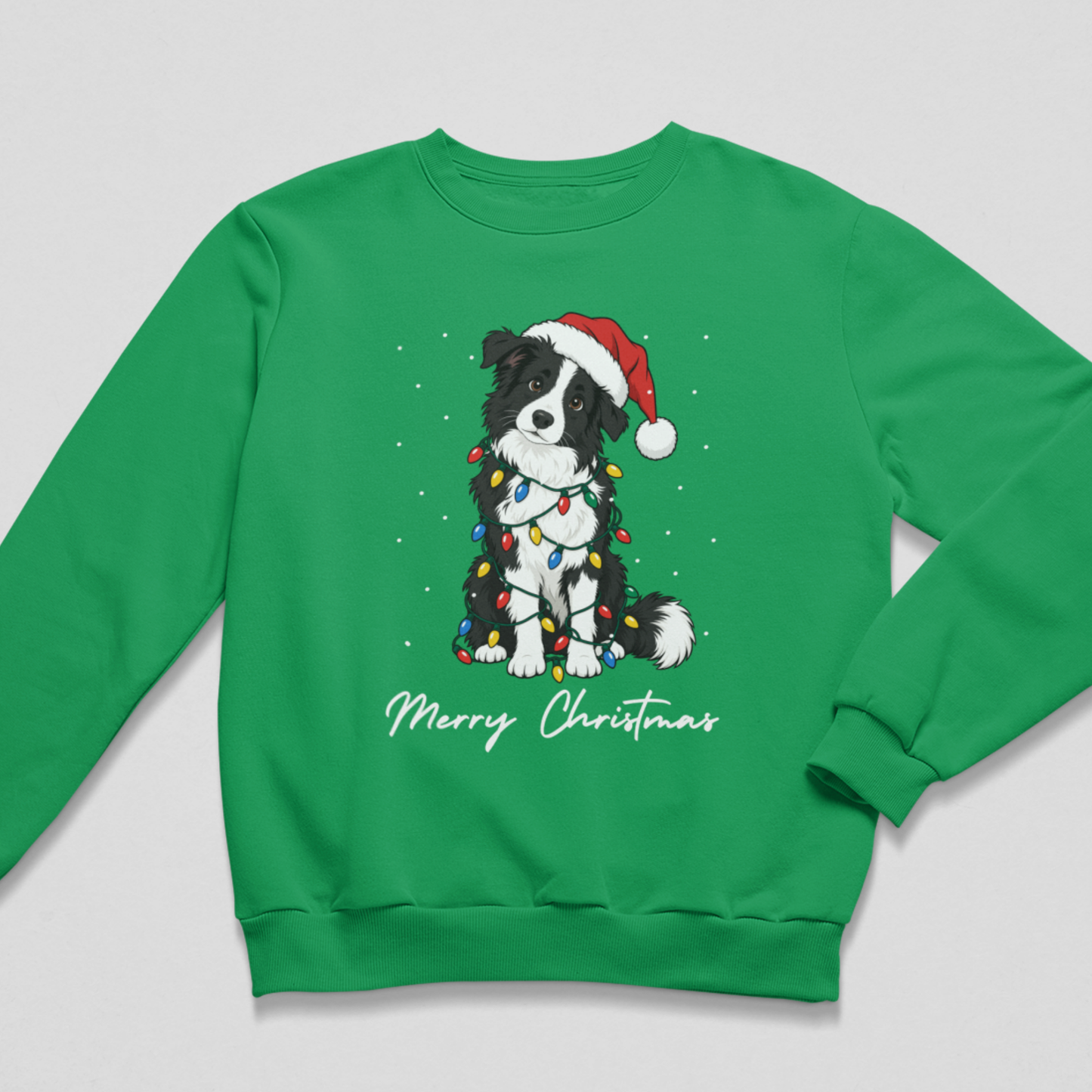 A bright kelly green children's holiday jumper laid flat against a neutral light background. The graphic showcases a cheerful Border Collie tangled in colourful fairy lights and a Santa hat, accented with "Merry Christmas" typography.