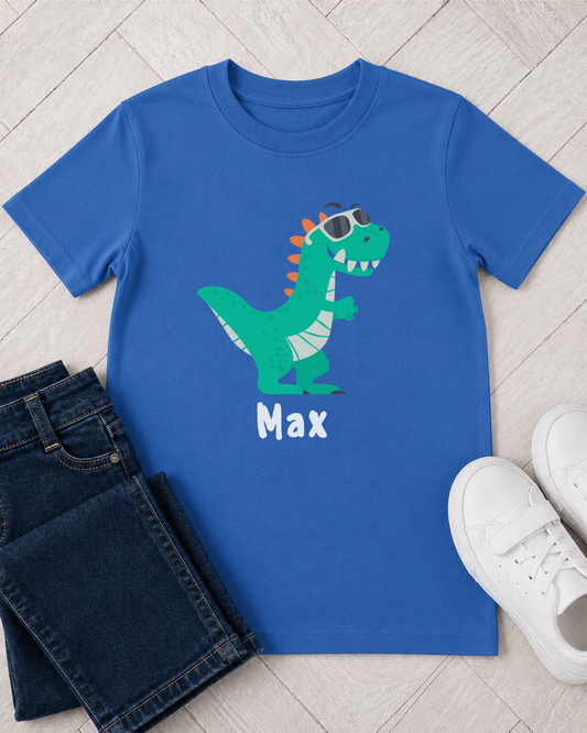 A kids royal blue short-sleeve kids' t-shirt featuring a green cartoon dinosaur wearing sunglasses with the name 'Max' printed below in white. The shirt is styled in a flat-lay arrangement with dark blue jeans, white sneakers, and a light wood floor background.