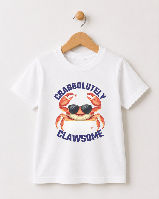 A white short-sleeve kids t-shirt hanging on a wooden hanger against a light beige wall. The shirt features a graphic of a smiling orange crab wearing black sunglasses, with the words 'CRABSOLUTELY' above and 'CLAWSOME' below in dark blue text.