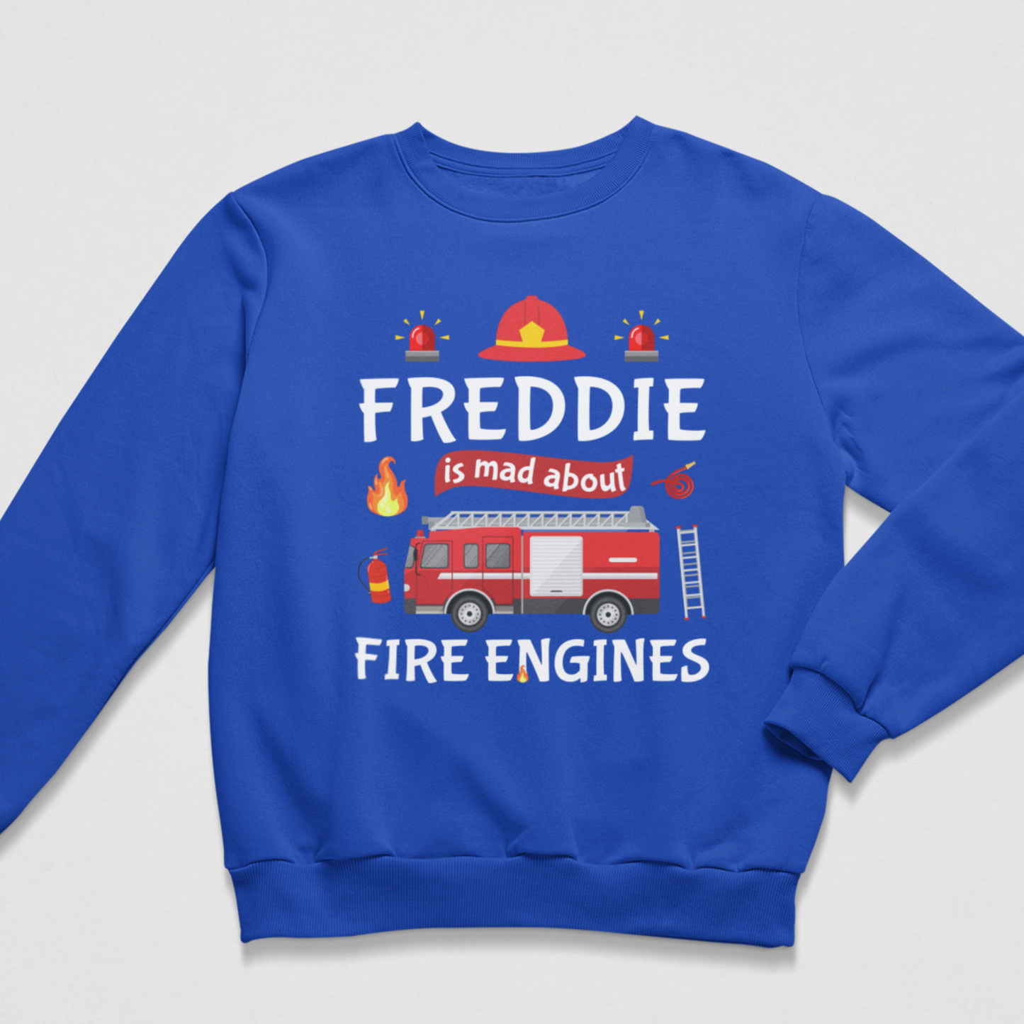 A boys royal blue crewneck sweatshirt featuring a graphic design on the front with the text 'FREDDIE is mad about FIRE ENGINES' and illustrations of a fire truck, helmet, alarms, and flames, displayed on a light grey background.