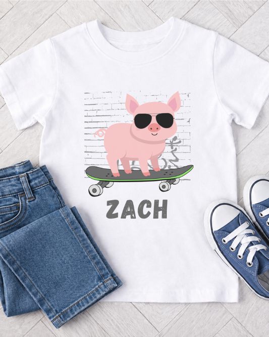 Kids white t-shirt with a pink pig on a skateboard graphic and 'ZACH' text, paired with blue jeans and shoes on a light wooden floor.