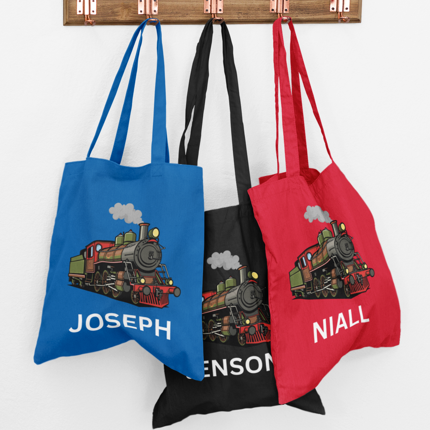 Three kids personalised steam train tote bags in red, royal blue and black hanging on hooks, each with a colourful steam engine and a child’s name printed underneath.
