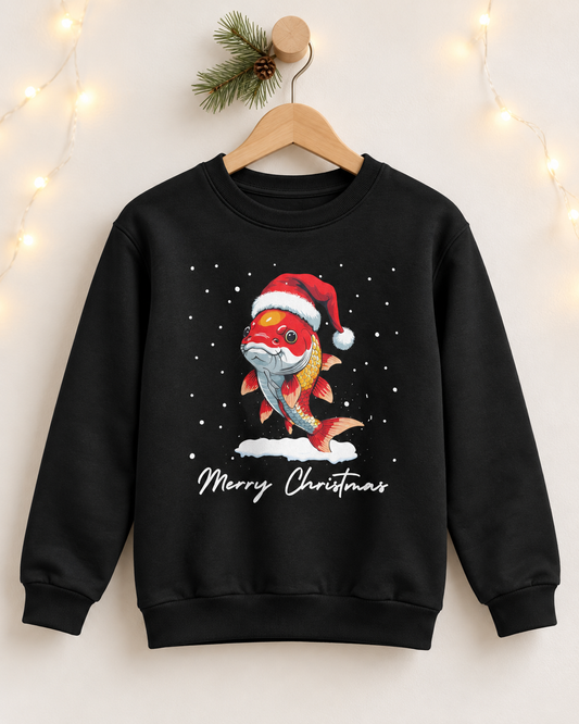 A black crewneck sweatshirt hanging on a wooden hanger against a warm beige wall with soft string lights. The sweatshirt features a graphic of a cartoon koi fish wearing a red Santa hat and white fur, standing on snow with 'Merry Christmas' written below in white script.