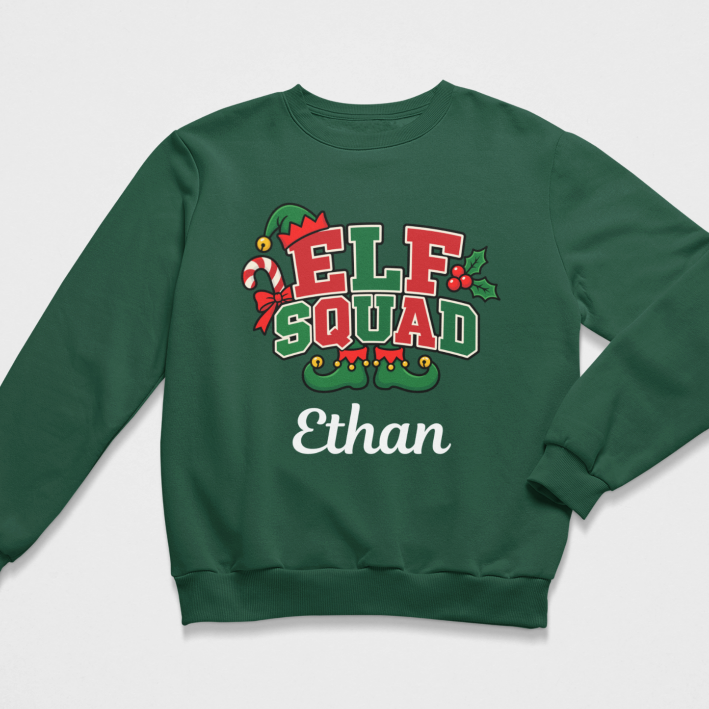 Flat lay of a dark green kids' Christmas crewneck sweatshirt featuring an "ELF SQUAD" holiday graphic with a candy cane and elf shoes, alongside the personalized name "Ethan