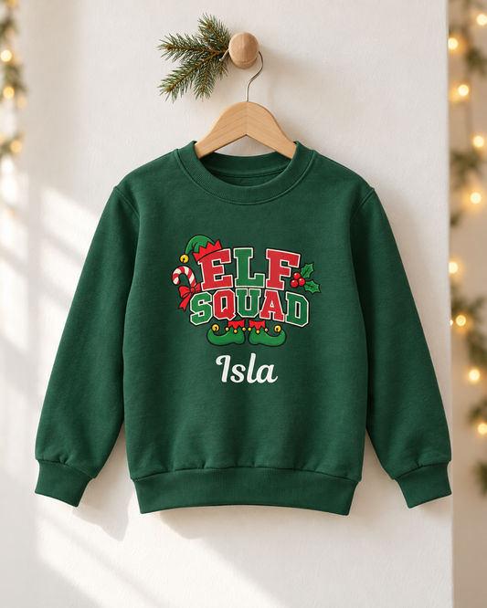 A dark green crewneck sweatshirt hanging on a wooden hanger against a white wall with holiday lights and pine branches. The sweatshirt features a festive graphic that reads 'ELF SQUAD' in red, green, and white with candy cane and holly details, and the name 'Isla' printed below in white.