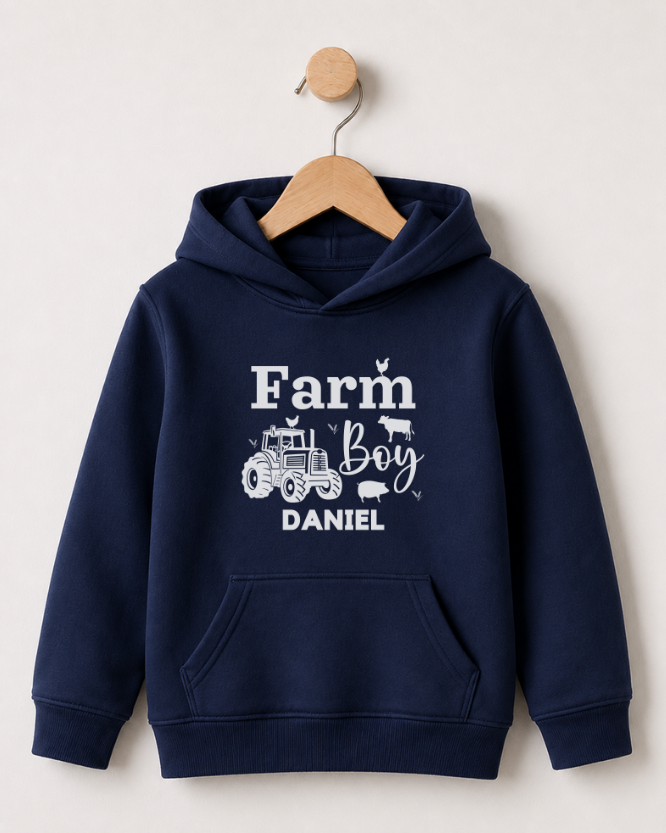 A navy blue kids pullover hoodie with a white graphic print reading 'Farm Boy DANIEL' and featuring a tractor, cow, and pig illustration, hanging on a wooden hanger against a light beige wall.