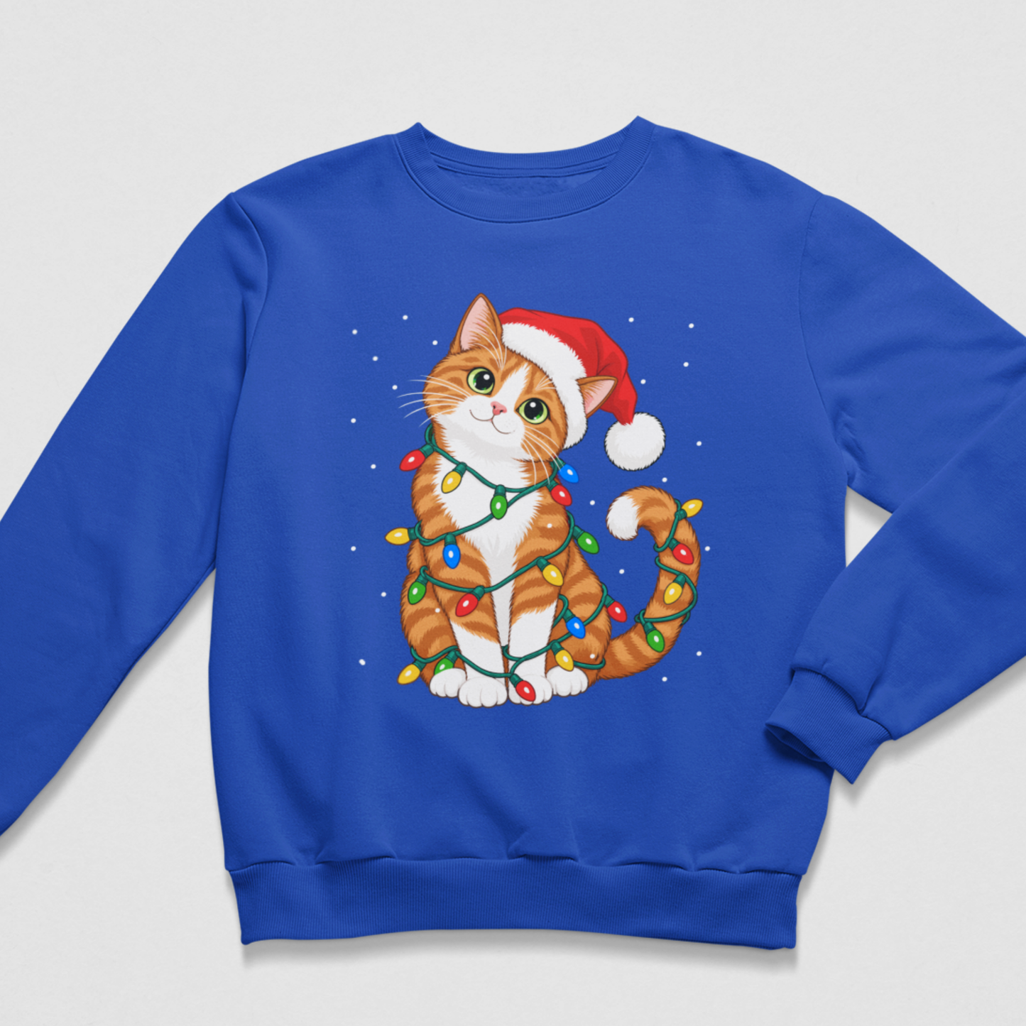 Flat lay of a royal blue kids' Christmas crewneck sweatshirt featuring an orange and white tabby cat wearing a Santa hat and wrapped in glowing festive lights, with delicate white snowflakes scattered around the design.