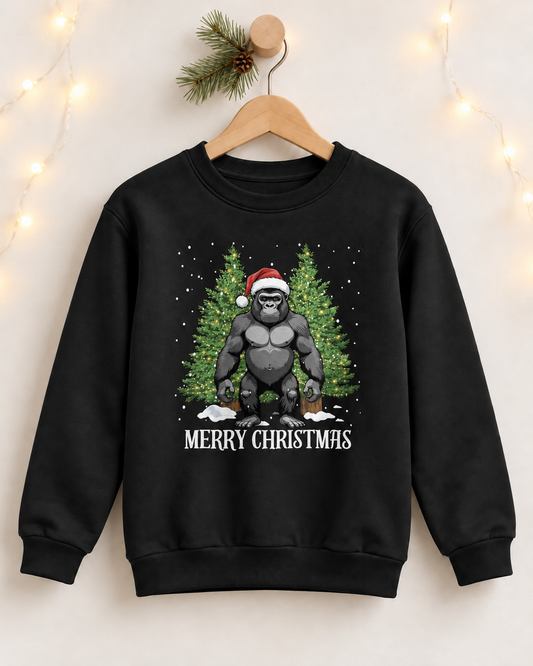 A kids black crewneck sweatshirt hanging on a wooden hanger against a warm, softly lit background with string lights. The sweatshirt features a graphic of a muscular gorilla wearing a Santa hat, standing in front of green Christmas trees with 'MERRY CHRISTMAS' printed below.