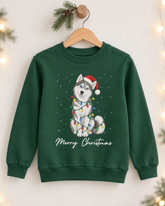 A kids dark green crewneck sweatshirt hanging on a wooden hanger against a warm off-white background with soft holiday lights and pine branches. The sweatshirt features a graphic of a grey and white husky wearing a red Santa hat and wrapped in colourful Christmas lights, with 'Merry Christmas' written below in white script.