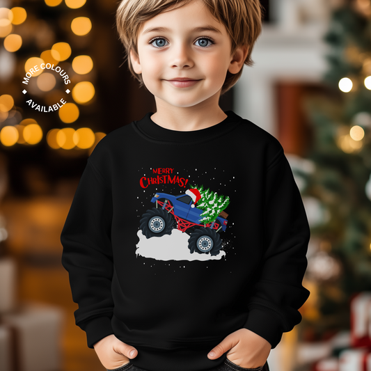 Smiling boy wearing a kids' Christmas monster truck sweatshirt with a festive tree on the back of a blue truck and “Merry Christmas” text, black jumper.