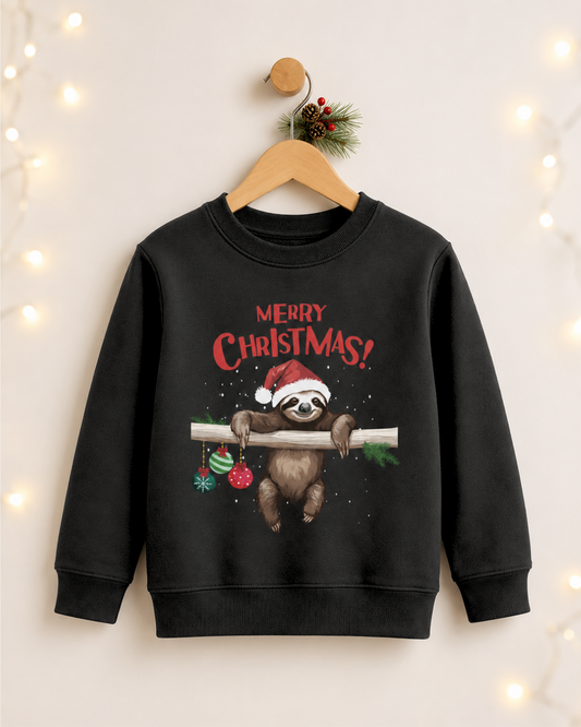 A black crewneck sweatshirt for kids featuring a Christmas graphic of a sloth wearing a Santa hat and hanging from a branch with 'MERRY CHRISTMAS!' text, displayed on a wooden hanger against a warm background with decorative fairy lights.