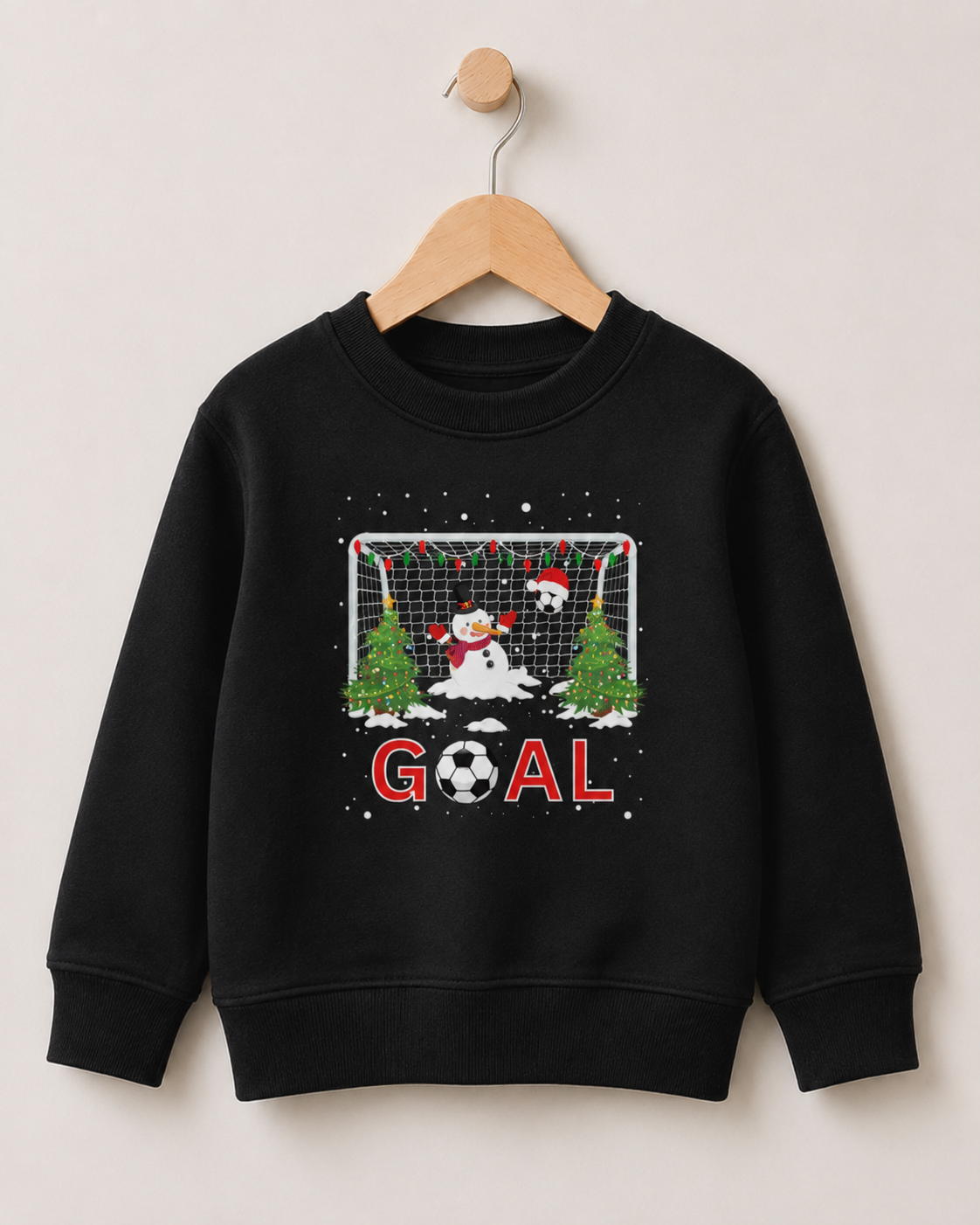 A kids black crewneck sweatshirt hanging on a wooden hanger against a light beige wall. The sweatshirt features a festive Christmas-themed soccer graphic with a snowman, a soccer ball, Christmas trees, a goal net, and the word 'GOAL' in red.