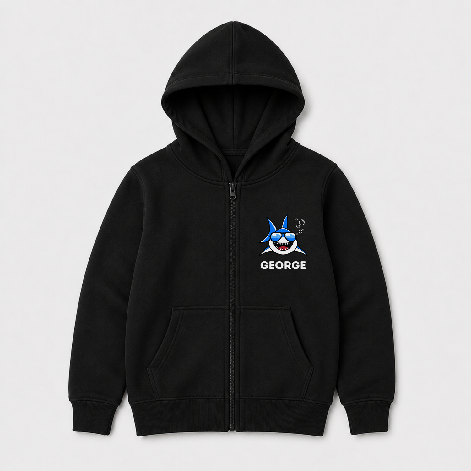 A kids black zip-up hoodie with a front kangaroo pocket, featuring a cartoon shark graphic and the name 'GEORGE' printed in white on the left chest, displayed on a light grey background.