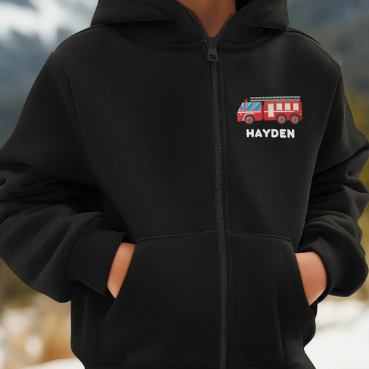 A close up image of a young boy wearing a black full zip hoodie with small printed motif of a red Fire Engine motif with the name "Hayden" underneath