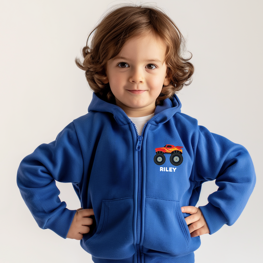 A little boy wearing a royal blue zip hoodie with a small monster truck with the name "Riley" printed underneath to chest area