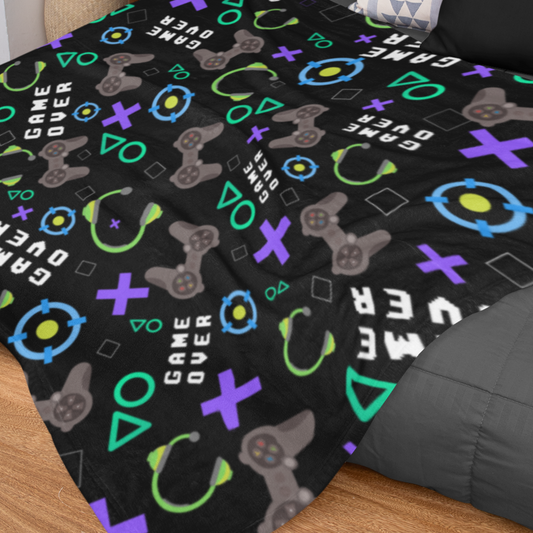 A gaming-themed throw blanket with various video game symbols and text patterns on a black background laid out on a bed.