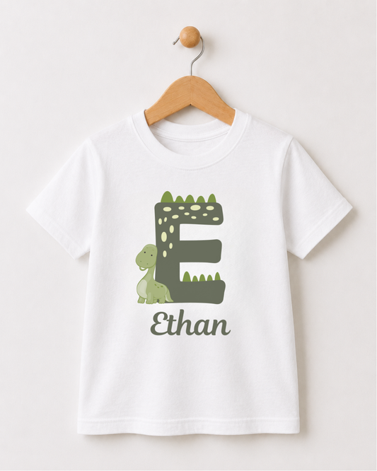 A white short-sleeve kids t-shirt hanging on a wooden hanger against a light beige wall. The shirt features a graphic of a green dinosaur sitting next to a large dinosaur themed letter 'E' with the name 'Ethan' printed below in a dark green font.