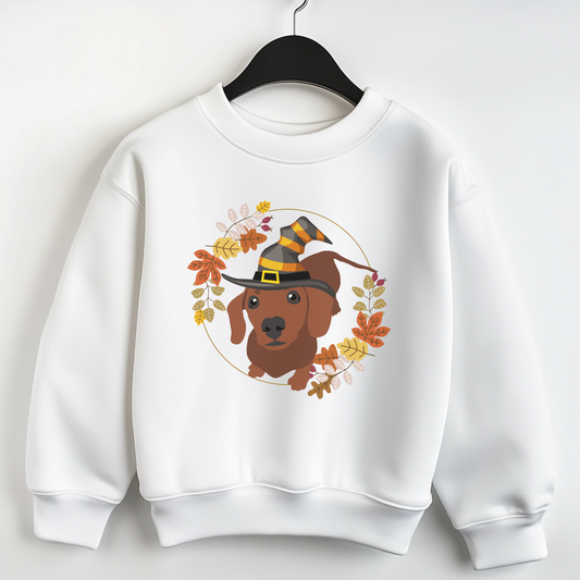 Kids white sweatshirt on a hanger featuring a cute dachshund wearing a witch hat, surrounded by autumn leaves in warm fall colours.